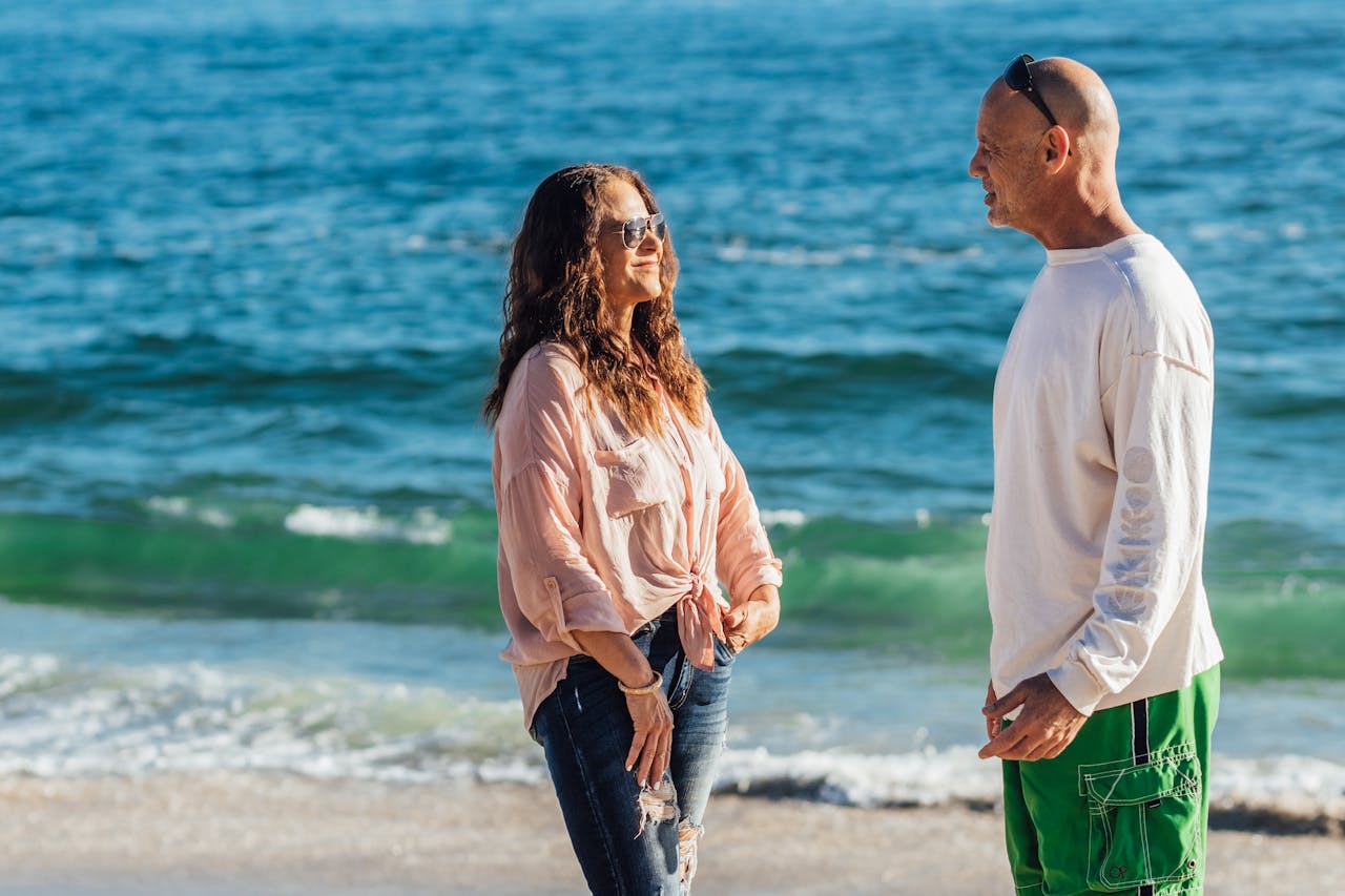 A couple enjoying a cheerful conversation on a sunny beach day, facing the ocean waves.